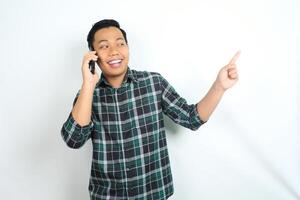 cheerful and carefree asian man wearing casual clothes pointing his hand while talking on cellphone, calling, with smiling face isolated on white background photo