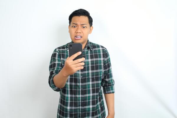 mad asian man holding cellphone and raising his hand looks angry and furious isolated on white background
