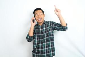 cheerful and carefree asian man wearing casual clothes pointing his hand while talking on cellphone, calling, with smiling face isolated on white background photo