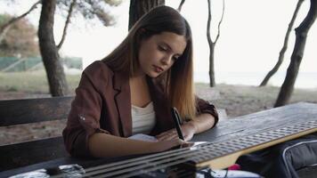 Girl Writes Music Notes On Sheet Music Sitting At The Park Table In The Nature video