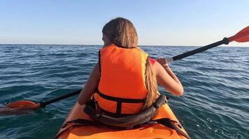 A woman in an orange life jacket paddles a kayak on a calm blue sea, with the setting sun on the horizon. Summer holiday vacation and travel concept video