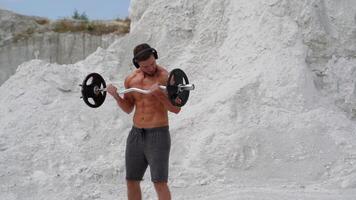 Bodybuilder lifting barbell and looking on his muscular body outside. Athlete man is training press muscles with metal barbell on white mountain background. video