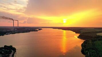 Beautiful sunset near the river in summer. Aerial view of evening environment of calm river among green fields and factory. Camera moves up. video