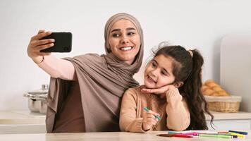 A mother and her daughter smile brightly as they take a selfie together. Colorful markers and crafting materials are spread across the table beside them, showcasing a fun, creative moment. video