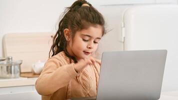 A cheerful girl sits at a kitchen table, focused on her laptop. She is wearing a cozy sweater and appears to be happily engaged with her work or activity, contributing to a lively atmosphere. video