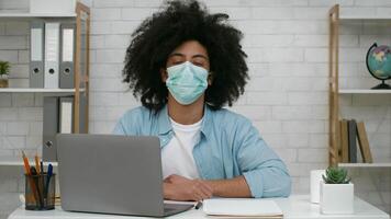 A young man sits at a desk with a laptop, wearing a mask and looking at the camera. He appears focused and engaged in an online class from a well-organized workspace. video