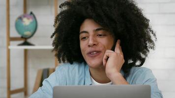 A young man with curly hair engages in a phone conversation, smiling while seated at a desk with a laptop in a well-lit room featuring a globe in the background. video