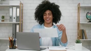 Black guy with curly hair is engaged in reviewing paperwork at a desk, using a laptop. The modern workspace features plants and organized materials, indicating focus and productivity. video