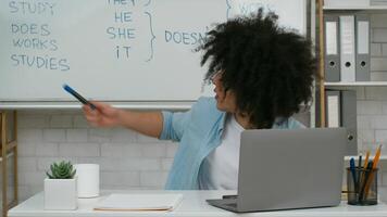 A young student with curly hair sits at a desk, focused on a laptop screen while studying English grammar concepts. A whiteboard in the background displays English pronouns and verbs. video
