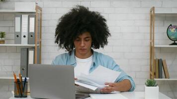 A young student sits at a desk, reviewing and organizing papers while working on a laptop in a contemporary study space with plants and bookshelves in the background. video