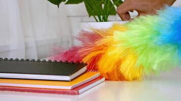 A hand reaches out to use a vibrant, rainbow-colored duster beside stacked notebooks and a green plant on a bright desk, emphasizing a moment of tidying up in a cheerful workspace. video