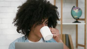 Black guy with curly hair sips coffee from a white mug while focused on his laptop. Sunlight illuminates the modern workspace, creating a calm atmosphere for productivity. video