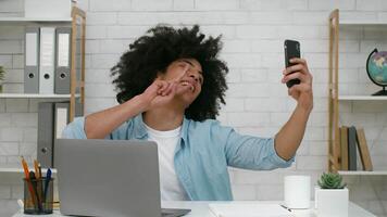 A young man with curly hair smiles and waves at his phone while sitting in his well-lit home office. He is surrounded by work essentials, creating a cheerful atmosphere for virtual interaction. video