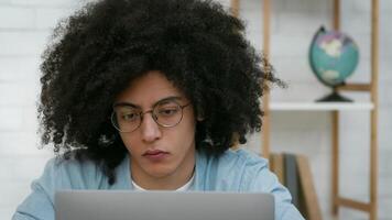 Black guy with thick curly hair and glasses is focused on his laptop in a stylish study room. Books and a globe are visible in the background, enhancing the scholarly atmosphere. video