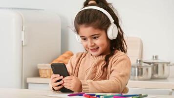 A girl sits at a table in a light kitchen, smiling as she looks at her smartphone. She wears headphones and has colorful markers nearby, suggesting a creative activity. video
