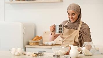 A young woman wearing a hijab measures ingredients in a bowl while baking in her bright, modern kitchen. Fresh bread is visible, showcasing a warm and inviting atmosphere. video