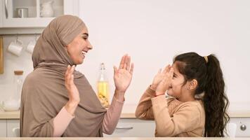 A mother wearing a headscarf and her daughter enjoy a playful clapping game together in a bright, contemporary kitchen filled with light. video