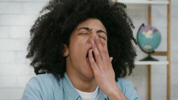 Black guy with voluminous curly hair appears to be yawning while seated at a desk in a cozy indoor setting, suggesting fatigue or boredom during a quiet moment. video