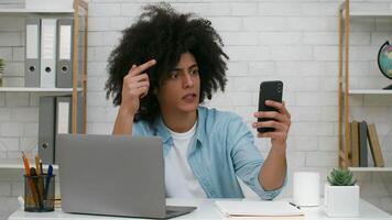 A young man with curly hair looks astonished while checking his smartphone. He is sitting at a desk with a laptop and notebooks, surrounded by a contemporary workspace. video