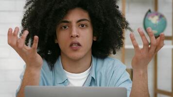 A young man with curly hair sits at a desk, looking confused and raising his hands in a questioning gesture while using his laptop. The setting is a cozy, well-lit indoor space. video