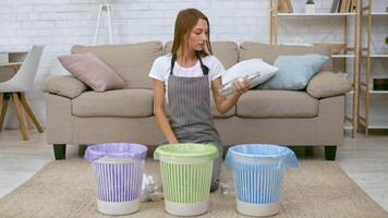 A woman is sorting waste items into three different color-coded bins in a bright living room. She carefully places recyclables in the appropriate container while sitting on the floor. video