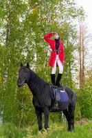Woman is standing on top of a black horse in a forest photo
