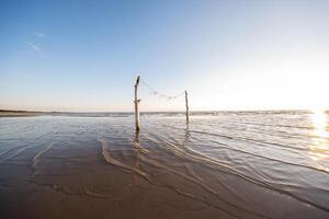 Close up of a serene beach with two wooden poles standing in the shallow water, adorned with string decorations under the warm light of sunrise. photo