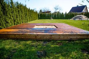 Freshly stained wooden deck with a central sandpit in a backyard surrounded by trees and bushes photo