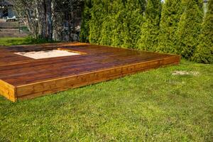 Freshly stained wooden deck with a central sandpit in a backyard surrounded by trees and bushes photo