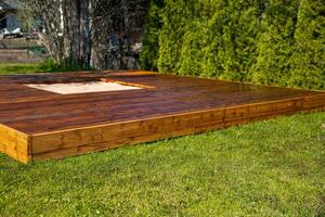 Freshly stained wooden deck with a central sandpit in a backyard surrounded by trees and bushes photo