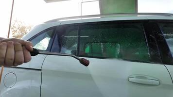 A person uses a pressure washer to clean a white car at a car wash, related to vehicle maintenance and spring cleaning video
