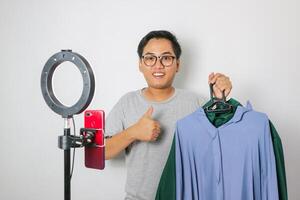 Excited young Asian men is holding up a pair of pants in front of a rack of clothes while showing thumb up gesture. Host live shopping concept. photo