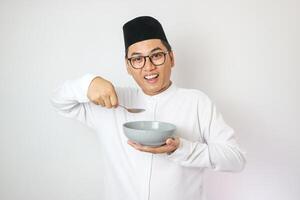 Excited young Asian Muslim man wearing Arabic clothes standing while holding bowl and saying yes it's time to break fast isolated on white background. People religious Islamic lifestyle concept. photo