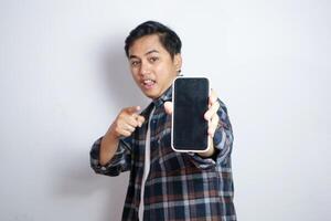Happy Asian man holding blank smartphone screen with happy and excited expression. Young Asian man wearing casual shirt smiling and showing white mobile phone screen for advertisement. photo