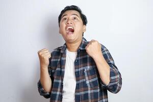A young Asian man in a casual shirt pumps his fists in excitement and smiles broadly against a solid yellow background. This image captures the essence of celebration and triumph photo