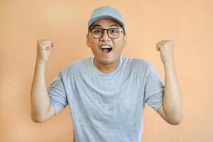 Excited and happy asian courier in grey uniform shouting yes and rejoicing, fist pump as celebrating great news, triumphing over white background. Delivery man looking euphoric and winning photo