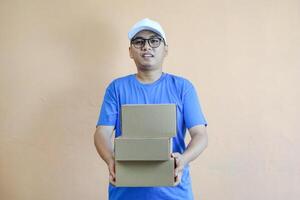 Happy Asian delivery man or courier is holding a cardboard box package parcel, smiling at the camera. Isolated on a orange background, Concept of satisfaction and efficiency in delivery services photo