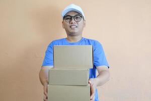 Happy Asian delivery man or courier is holding a cardboard box package parcel, smiling at the camera. Isolated on a orange background, Concept of satisfaction and efficiency in delivery services photo