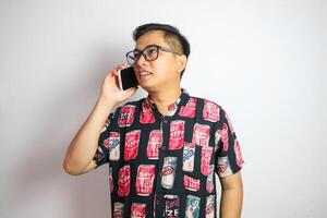 A fun, excited Asian man tourist in summer clothes and sunglasses is talking on the phone with someone, posing with a luggage on an isolated white background. summer vacation, beach trip, journey photo