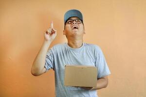 Asian delivery man is pointing at empty copy space while holding a cardboard box package parcel, Isolated on a white background, advertisements, promotions, or announcements, delivery services photo