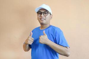 smiling delivery man in blue uniform carrying packages while gesturing thumb up sign isolated on orange background photo