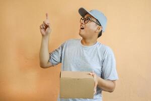 Asian delivery man is pointing at empty copy space while holding a cardboard box package parcel, Isolated on a white background, advertisements, promotions, or announcements, delivery services photo