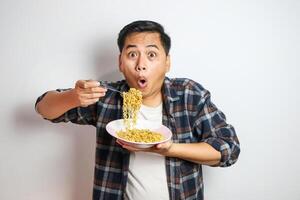 An Asian man in a casual shirt eagerly eats noodles from a plate, his expression filled with delight. The plain white background focuses on his enjoyable dining experience. photo