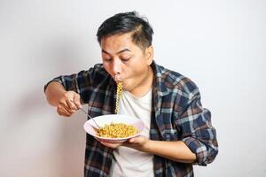 An Asian man in a casual shirt eagerly eats noodles from a plate, his expression filled with delight. The plain white background focuses on his enjoyable dining experience. photo