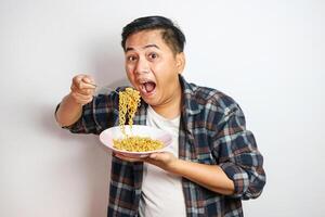 An Asian man in a casual shirt eagerly eats noodles from a plate, his expression filled with delight. The plain white background focuses on his enjoyable dining experience. photo