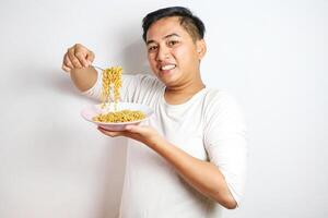 A happy, euphoric Asian young man in a tshirt eating instant fried noodles using fork isolated over a white background. stylish asian man with peek a boo hair eats instant noodle photo