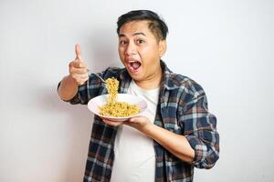 Young Asian man in a white t-shirt enthusiastically eating instant noodles from a bowl with a fork. Studio shot on a plain background, highlighting his joyful expression and casual fashion photo