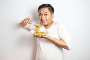 Young Asian man in a white t-shirt enthusiastically eating instant noodles from a bowl with a fork. Studio shot on a plain background, highlighting his joyful expression and casual fashion photo