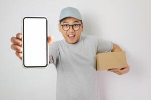 Delivery Service Application. Smiling indonesian man showing smartphone with blank screen for mockup, wearing red cap, uniform and holding cardboard box,courier guy posing on white background photo