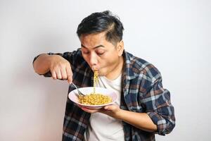 An Asian man in a casual shirt eagerly eats noodles from a plate, his expression filled with delight. The plain white background focuses on his enjoyable dining experience. photo
